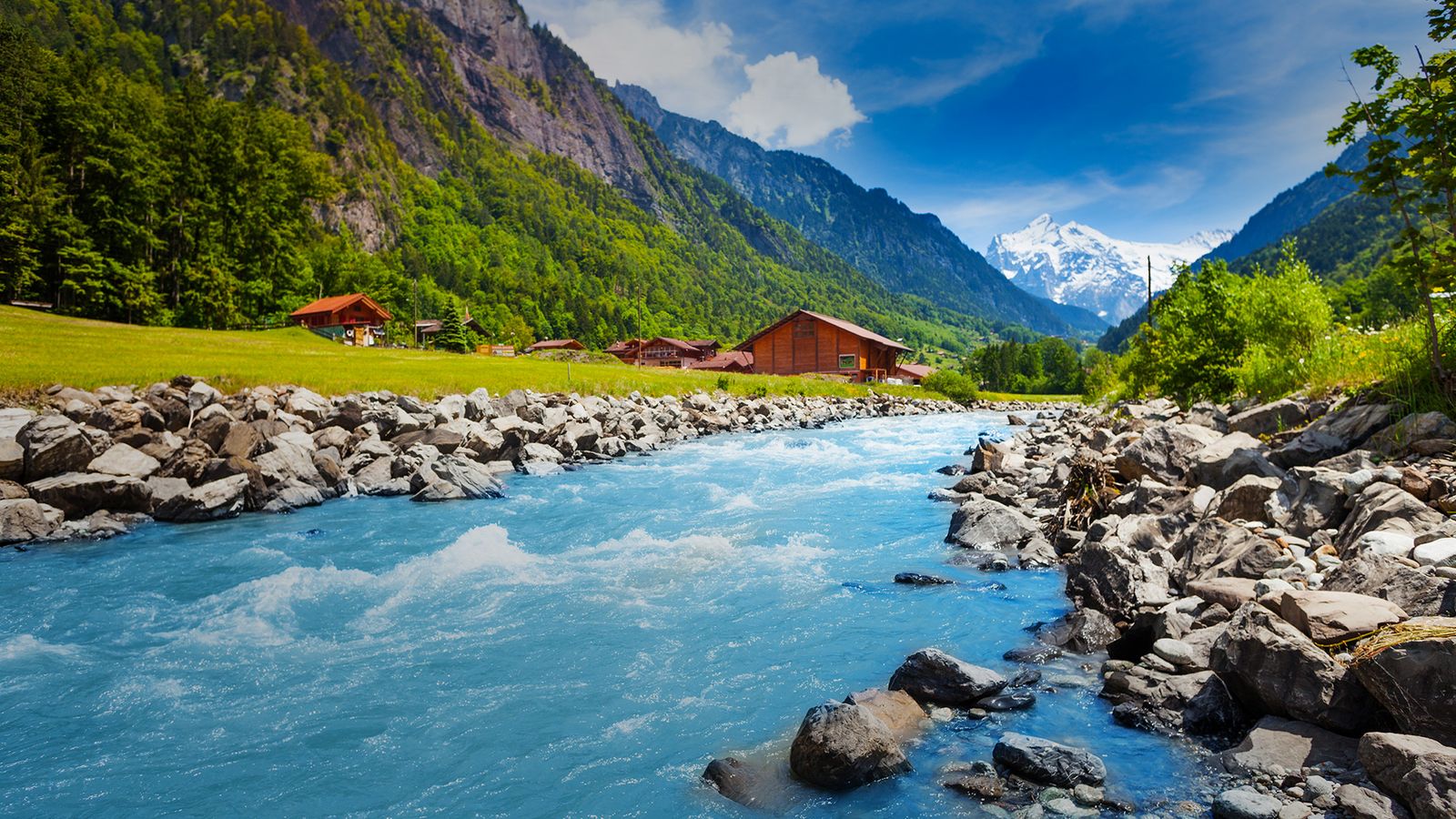 Ein türkisfarbener, klarer Bergfluss fließt durch eine alpine Landschaft mit felsigen Ufern. Im Hintergrund sind grüne Wiesen, vereinzelte Holzhäuser sowie bewaldete Berge mit einem schneebedeckten Gipfel unter blauem Himmel zu sehen.