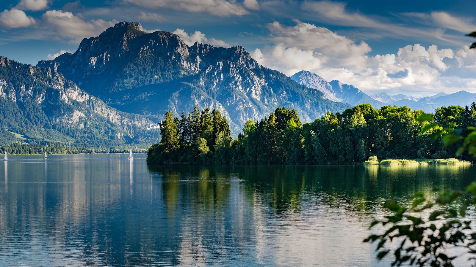 Ein stiller See spiegelt die umliegenden Berge und den teils bewölkten Himmel wider. Am Ufer verläuft eine dichte Reihe grüner Bäume, während in der Ferne mehrere Segelboote über das ruhige Wasser gleiten und eine friedliche Stimmung erzeugen.